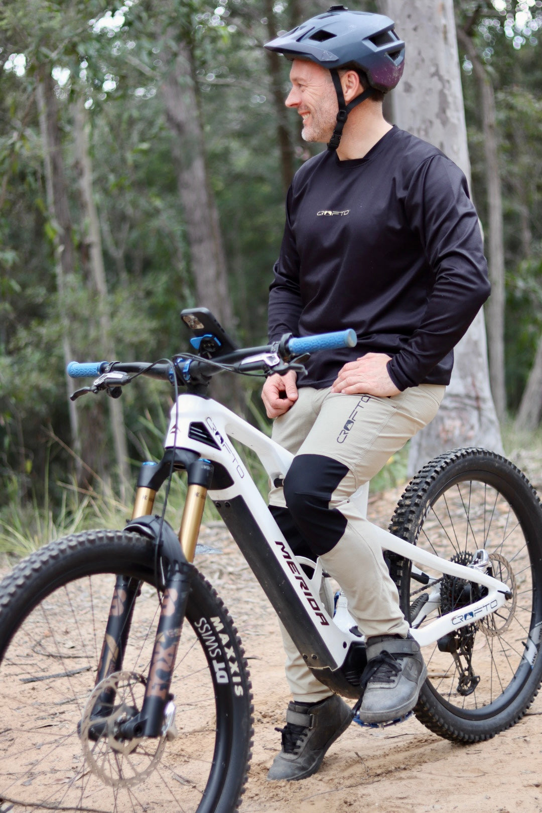Rider wearing CROFTO Amaroo MTB pants in sand colour and CROFTO enduro mtb jersey sitting on a mountain bike on a dirt trail.