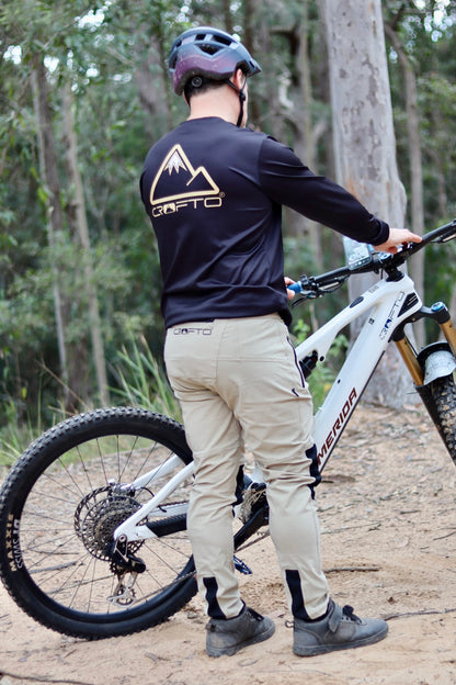 Rider wearing CROFTO Amaroo MTB pants in sand colour and CROFTO enduro jersey in black standing next to a mountain bike on a dirt trail.