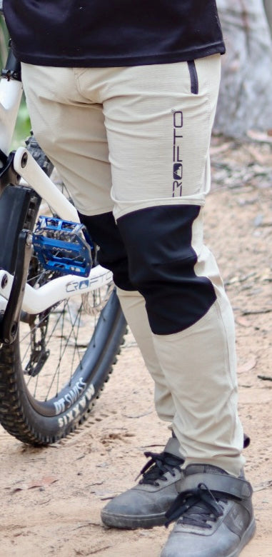 Rider wearing CROFTO Amaroo MTB pants in sand colour beside a mountain bike on a dirt trail.
