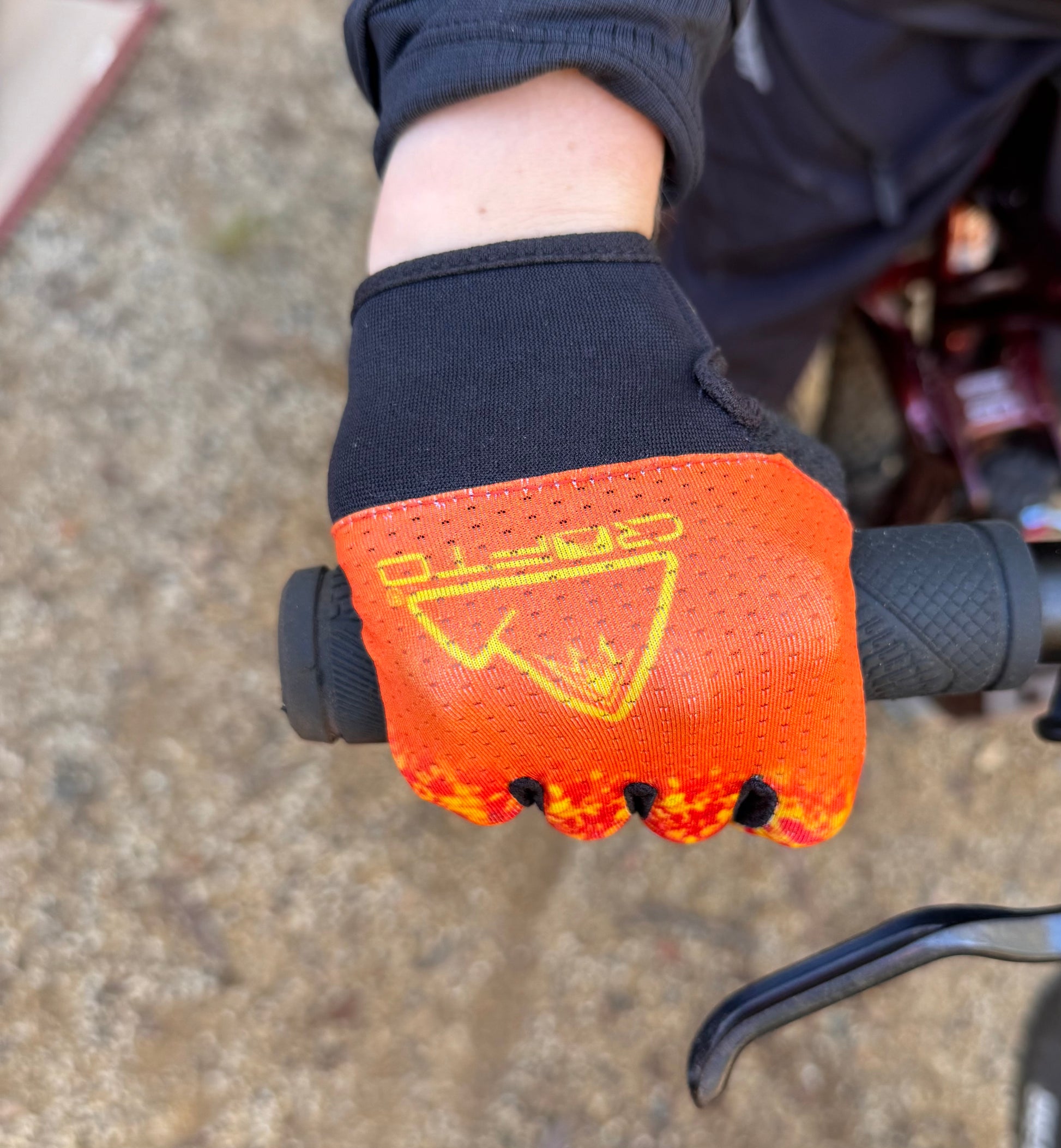 A close-up of a hand wearing a crofto apex pro kids MTB glove with a red, orange, and yellow flame-like pattern, gripping a black bicycle handlebar on an outdoor dirt trail at Ourimbah mtb park