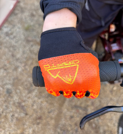 A close-up of a hand wearing a crofto apex pro kids MTB glove with a red, orange, and yellow flame-like pattern, gripping a black bicycle handlebar on an outdoor dirt trail at Ourimbah mtb park