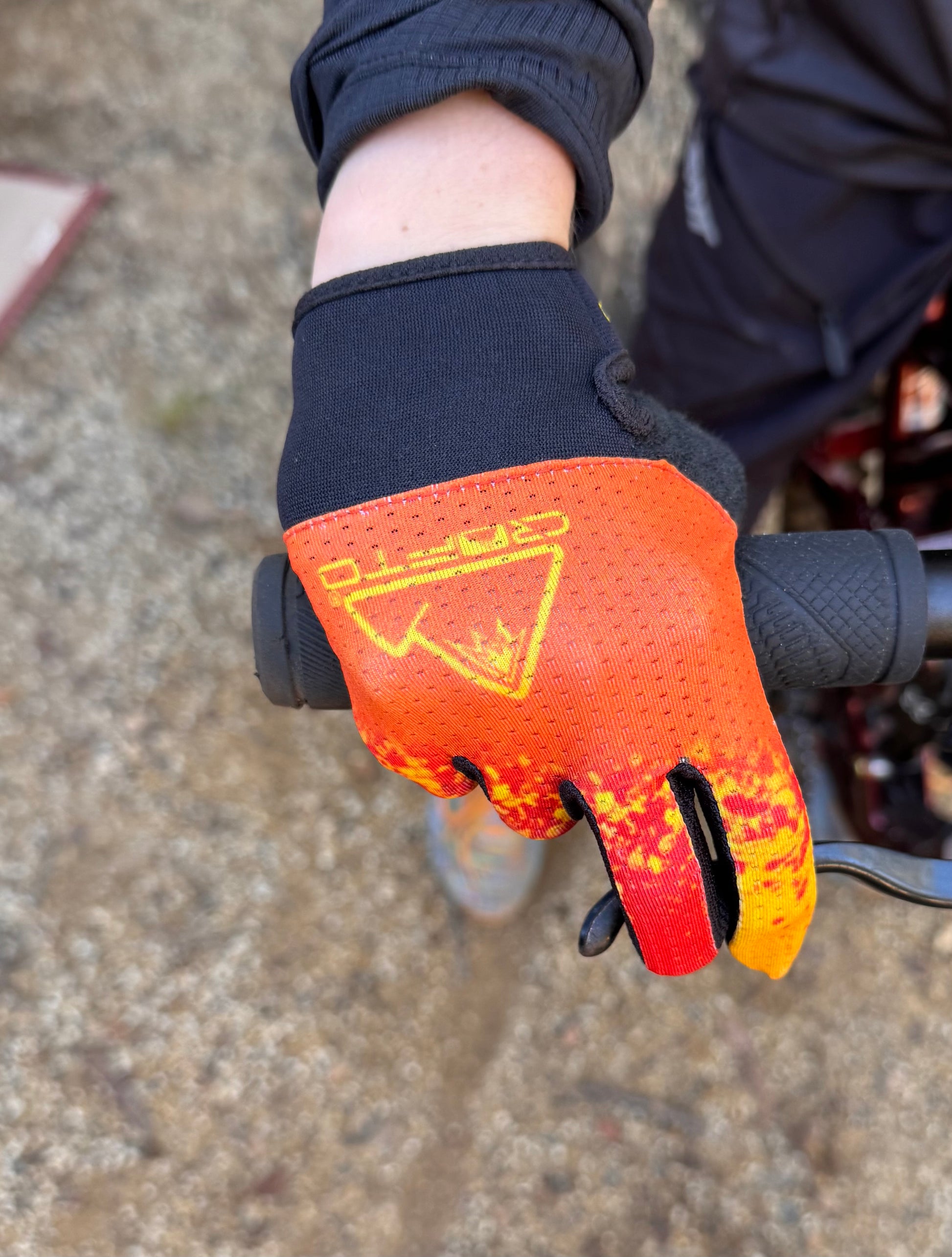 A close-up of a hand wearing a crofto apex pro kids MTB glove with a red, orange, and yellow flame-like pattern, gripping a black bicycle handlebar on an outdoor dirt trail at Ourimbah mtb park