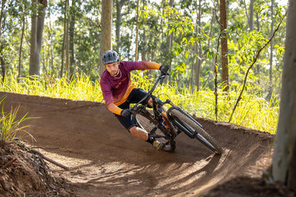 A mountain biker wearing a maroon and gold crofto jersey, navy crofto jorts and a helmet leans sharply into a dirt berm on a forest track.