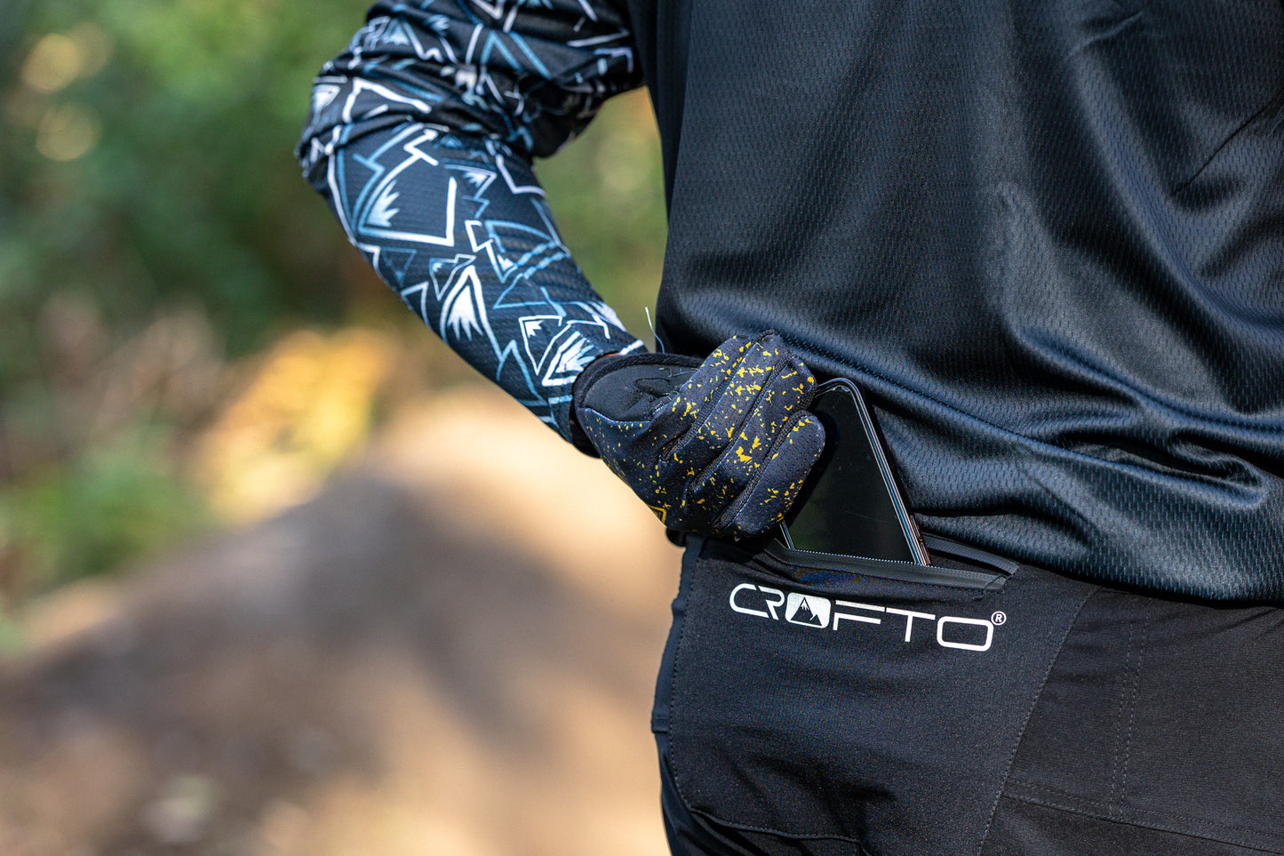 Close-up of a person outdoors wearing a black long-sleeve crofto mozaic jersey and black amaroo pants putting a phone in the rear pocket