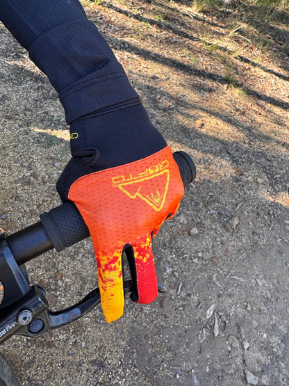 A close-up of a hand wearing a crofto apex pro kids MTB glove with a red, orange, and yellow flame-like pattern, gripping a black bicycle handlebar on an outdoor dirt trail.