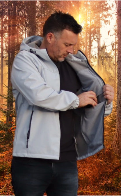 Man wearing a light gray crofto soft shell jacket  with it open and putting a phone in the inside pocket in a forest during sunrise.