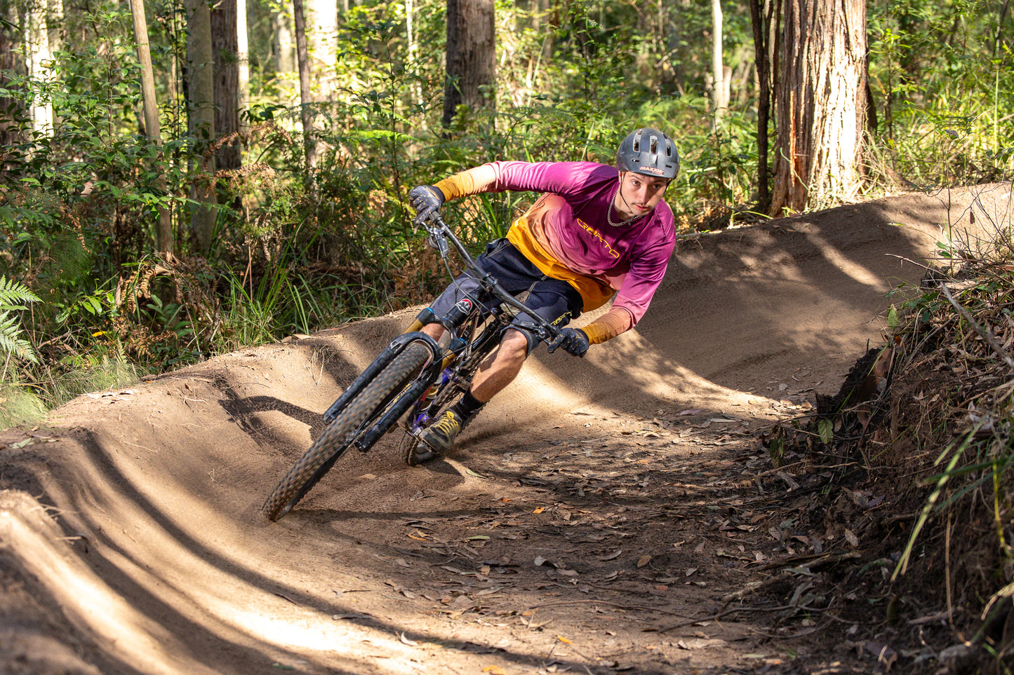 A mountain biker wearing a maroon and gold crofto jersey, navy crofto jorts and a helmet leans sharply into a dirt berm on a forest track.