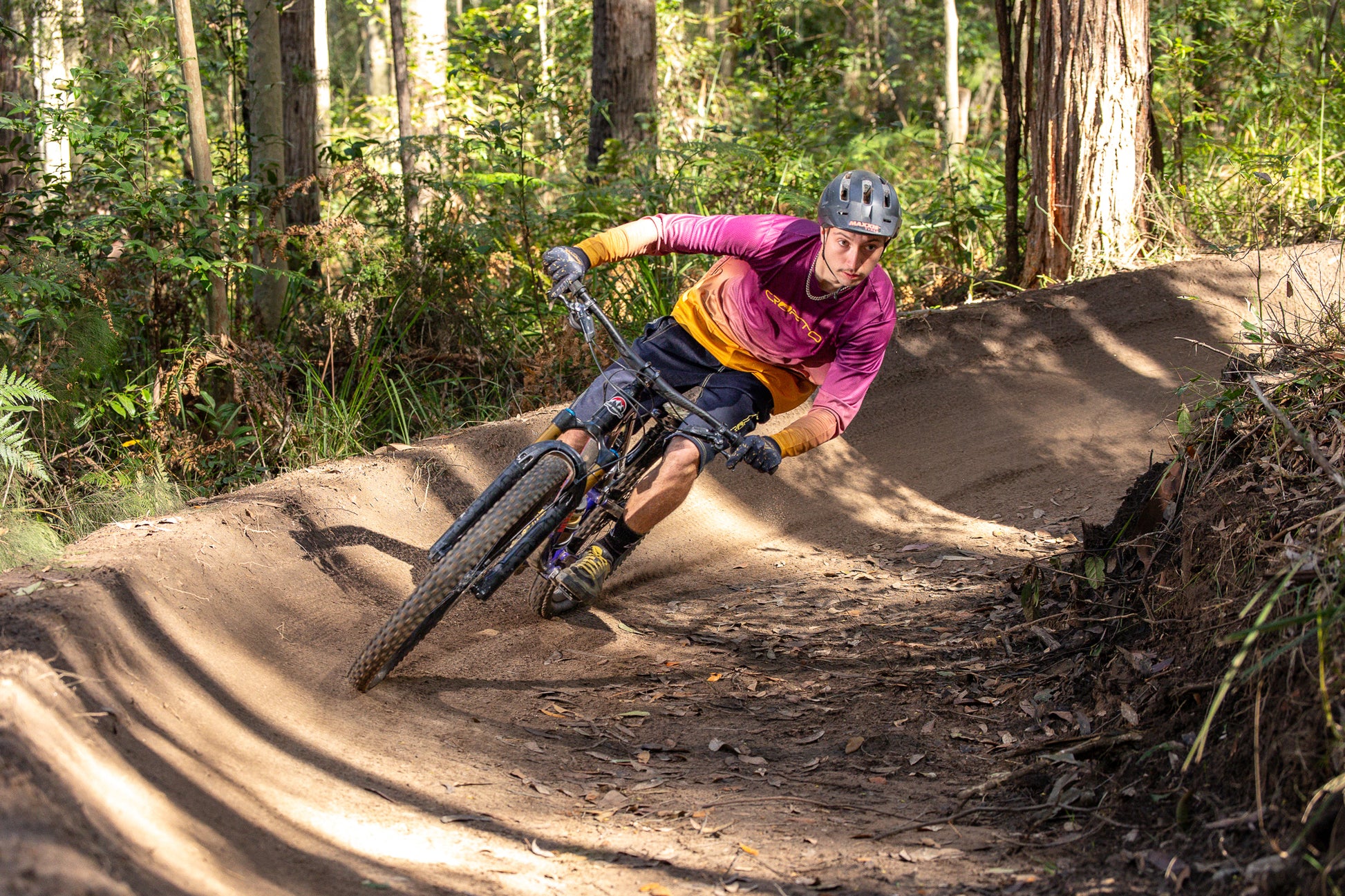 A mountain biker wearing a maroon and gold crofto jersey, navy crofto jorts and a helmet leans sharply into a dirt berm on a forest track.