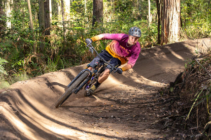 A mountain biker wearing a maroon and gold crofto jersey, navy crofto jorts and a helmet leans sharply into a dirt berm on a forest track.