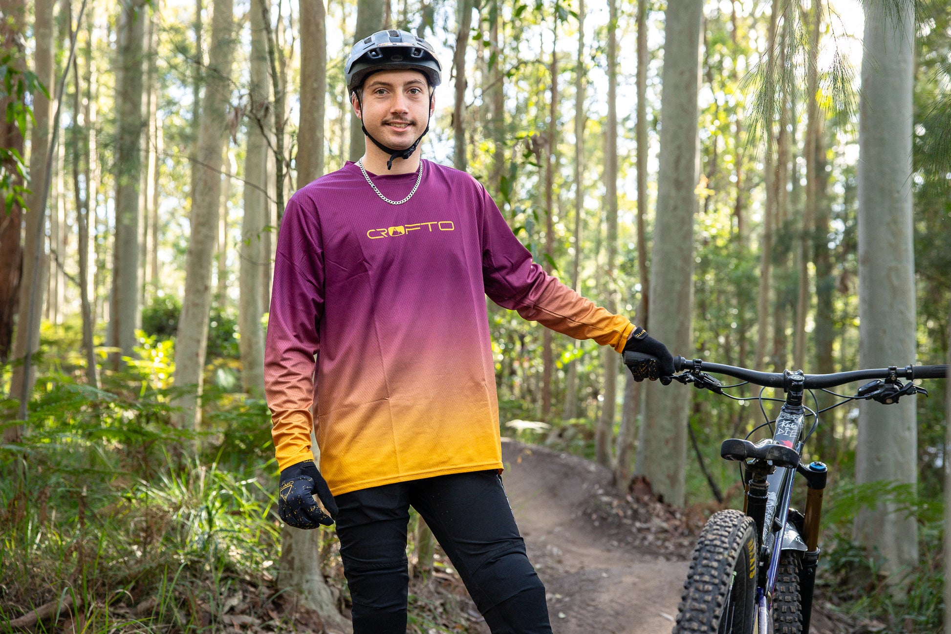 Man standing next to a mountain bike in a forest wearing a CROFTO Fade riding jersey and Amaroo MTB pants