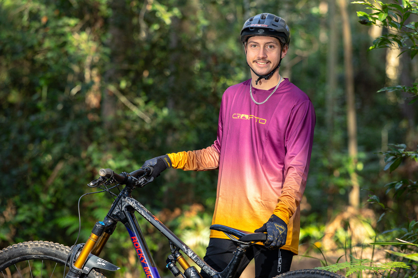 A man wearing a mountain bike helmet and a CROFTO fade long sleeve MTB jersey in a forest next to his bike