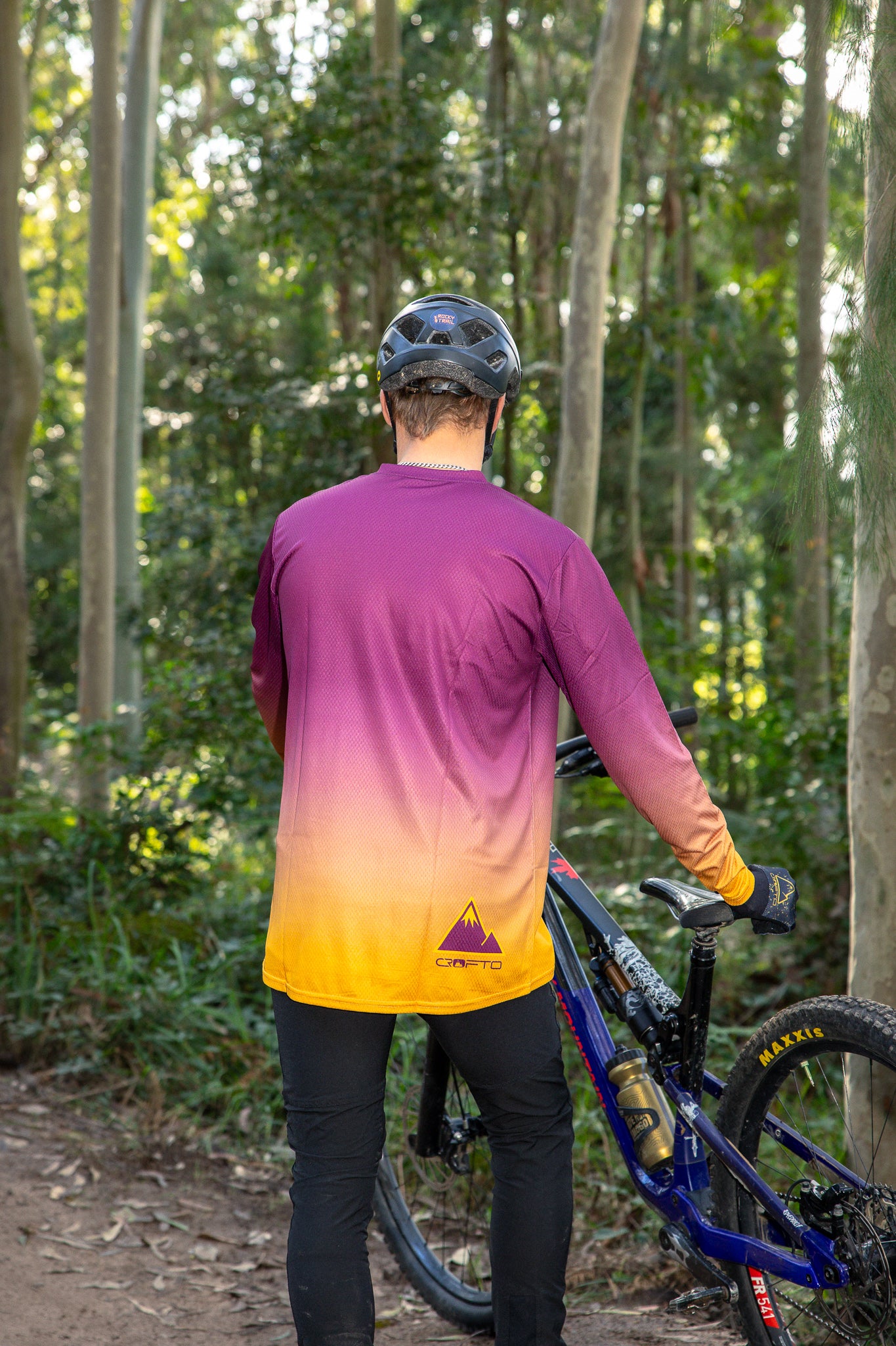 a back view of a man wearing a mountain bike helmet and a CROFTO fade long sleeve MTB jersey in a forest next to his bike