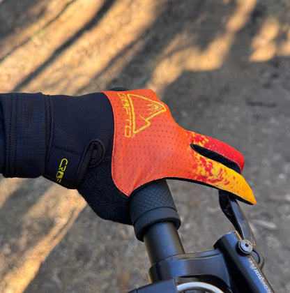 A close-up of a hand wearing a crofto apex pro kids MTB glove with a red, orange, and yellow flame-like pattern, gripping a black bicycle handlebar on an outdoor dirt trail at Ourimbah mtb park